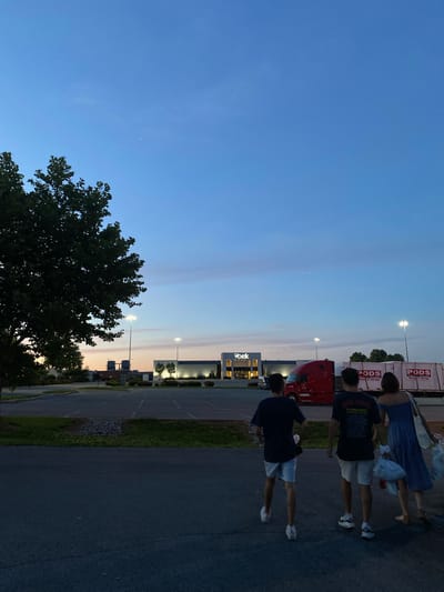 At dusk, three individuals walk away from the camera on a paved surface in front of a commercial building. The building, a Belk department store, is illuminated and features signage. To the right of the building, a red semi-truck is parked next to a PODS moving and storage container. A large tree with dark green foliage is positioned on the left side of the frame, casting a shadow. The sky above is a gradient of deep blue and lighter hues, with faint traces of pink and orange near the horizon. The parking lot in front of the building appears mostly empty, with visible parking lines. The overall environment suggests a retail area in a suburban or semi-rural setting within the city of Halfway, United States of America.