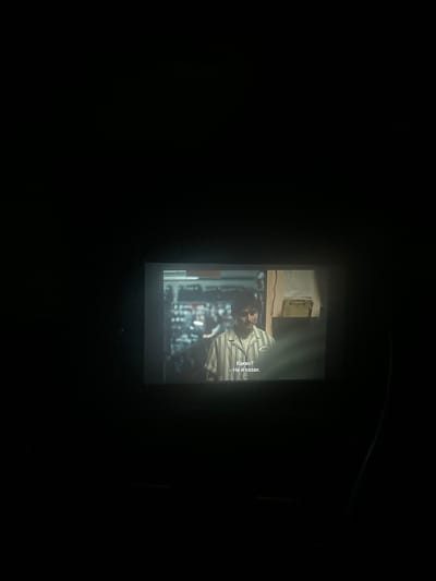 The image shows a projected movie scene in a dark room. The projected image displays a man wearing a striped shirt. Text is visible at the bottom left of the projected image, which reads "Какво? - Не и казах." This translates to "What? - And I didn't say." The scene appears to be indoors, possibly a store or office, with shelves and items visible in the background. The overall environment is dark, with the projection being the main source of light. There is no identifiable information linking this scene to the city of Byala Slatina, Bulgaria.