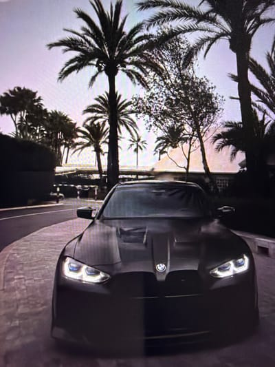 The image shows the front of a dark-colored car, likely a sports car, parked outdoors. The car's headlights are on, casting a bright, reflective glow. In the background, silhouetted palm trees and foliage are visible against a light-colored sky, suggesting a tropical or subtropical setting, possibly during sunrise or sunset due to the soft lighting. The car is positioned on a paved surface that appears to be a road or walkway, with a textured wall or structure to its right. The image is oriented vertically, and the car's front is angled towards the viewer. There is no discernible text within the image. The overall impression is one of a scenic, possibly exotic, location.