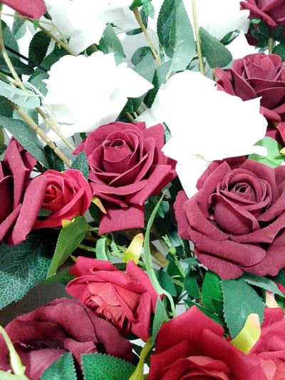 The image is a close-up, overhead view of a bouquet of artificial red and white roses interspersed with green leaves and thin brown stems. The roses are clustered together, with some in full bloom and others partially closed. The lighting is bright, causing some areas to be overexposed, particularly the white roses. There are no people or any discernible elements of the city of Shirin, Uzbekistan present in the image.