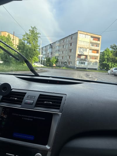 The image depicts a view from inside a car, likely stopped on a street in Peresvet, Russia. Through the windshield, a rainbow can be seen arching over a multi-story residential building typical of Soviet-era construction. Green trees surround the building. On the car's dashboard, the radio screen displays a message in Russian. Windshield wipers are visible.