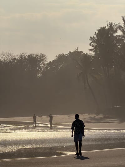 The image depicts a coastal scene in Selemadeg, Indonesia, at either sunrise or sunset, characterized by strong backlighting and a hazy atmosphere. The sky occupies the upper portion, exhibiting a pale, diffused light with subtle cloud formations.

The background features a dense, dark expanse of tropical vegetation, primarily silhouetted trees on the left and prominent palm trees on the right, suggesting a thick jungle or coastal forest bordering the beach. The sun's position behind these trees creates a strong lens flare effect and diffuses light through the foliage, obscuring specific details of the vegetation.

In the mid-ground, a body of water, likely the ocean, stretches across the frame. The surface of the water reflects intense specular highlights from the low sun, creating a shimmering, golden effect. Patches of wet sand are visible within and adjacent to the water. Two individuals are present in the mid-ground, standing partially submerged in the water or on a wet sandbar. They are entirely silhouetted, with their forms indistinct, and appear to be wearing shorts.

The foreground is dominated by a stretch of wet sand. A single male individual walks from the right side of the frame towards the left, facing generally towards the viewer but with his head turned slightly left. He is silhouetted against the bright water and sky, wearing a short-sleeved, collared shirt with an indiscernible pattern, shorts, and shoes. He casts a distinct shadow extending forward from his feet. The overall scene is peaceful and characterized by the dramatic effects of the low-angle sunlight.