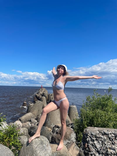 A blonde woman in a white bikini and white hat stands on a rocky breakwater with her arms outstretched. The breakwater is made of large concrete blocks and natural stones. The sea is dark and choppy, with a clear blue sky dotted with white clouds above. In the distance, a red buoy and some small white boats are visible. Lush green foliage grows on the right side of the breakwater. The scene appears to be the shoreline of Lake Beloye in Belozersk, Russia, known for its historic white-stone architecture and natural beauty.