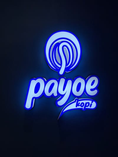 This media file features a neon sign with a blue and white color scheme against a dark, textured background. The sign displays two distinct elements. On the left, there's a stylized, circular logo resembling a coiled rope or a stylized 'E'. On the right, the word "Payoe" is written vertically in a script font, with "kopi" written above and to the right of "Payoe," also in a script font and oriented diagonally. The neon lights of the sign cast a glow on the dark surface, creating a subtle luminous effect. The overall aesthetic is modern and likely intended for display in a commercial setting, possibly a cafe or shop, given the word "kopi" (Indonesian for coffee). The setting appears to be indoors, possibly at night, due to the prominence of the neon lighting and the dark backdrop. There are no people or specific environmental cues to indicate time of day or weather. The image focuses solely on the illuminated sign.