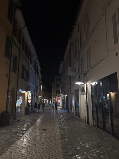 The image depicts a narrow, cobblestone street in Ravenna, Italy, at night. The street is lined with buildings featuring shops on the ground floor and windows with shutters on the upper levels. Lampposts illuminate the scene, casting light on the wet cobblestones, suggesting recent rain. Several people are walking down the street. On the left, a building facade is painted yellow, and a shop window displays merchandise. On the right, buildings are painted white, with large glass shopfronts. A clock is visible on one of the white buildings. The sky is dark, indicating nighttime.
