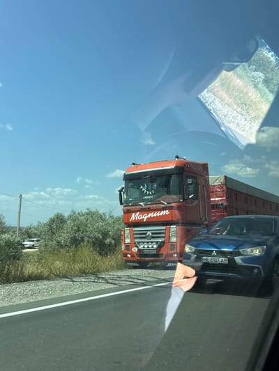 In Kobleve, Ukraine, on a sunny day, a blue Mitsubishi SUV is captured in traffic next to a red Renault Magnum semi-truck. The SUV's license plate reads "HE 2107 AB". The semi-truck has "Magnum" written on its side. The scene unfolds on a paved road bordered by a gravel shoulder and sparse vegetation, with a clear blue sky dotted with fluffy white clouds above. The image is taken from inside another vehicle, evidenced by reflections on the windshield.