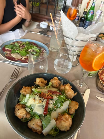 The image shows a table at a restaurant with various foods and drinks. The table is covered in a beige tablecloth and has a dark brown wooden surface underneath. The food items include a salad with lettuce, fried bread, parmesan cheese, and other seasonings in a black bowl, a plate with a meat salad covered in greens, and a pizza slice. The drinks include an orange drink in a stemmed glass, a glass of water, a tall glass with a straw containing a clear beverage, and another glass of water with a straw in it. There is also an assortment of empty liquor bottles and glasses on the table. The restaurant is located in the city of Santa Ana, Costa Rica, at the coordinates 9.934 latitude and -84.179 longitude. A person can be seen seated at the table with their hands clasped in their lap. The person appears to be wearing a black shirt and a white skirt. They have their back turned to the camera. They are not interacting with the food on the table. There are other people out of view, but they are not visible in the image. The image was taken indoors. The lighting in the photo is warm and natural. There are many elements present in the scene and the image is a very simple, yet detailed, presentation of a casual dining experience.