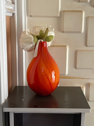 A close-up shot features an orange, tear-shaped glass vase adorned with artificial white flowers. The vase is suspended near a dark brown cabinet door. The background consists of a textured white wall with square-shaped protrusions and a white windowsill with ridges. A thin, straw-like object extends vertically behind the vase. The location within Reifild-Ist, Nigeria, is not determinable based on the image.