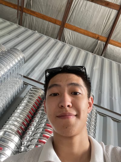 A young man with short dark hair, wearing sunglasses perched on his head and a white polo shirt, is pictured from the chest up, looking directly at the camera. He is indoors, with a corrugated metal wall behind him and a ceiling of wooden beams and reflective insulation visible above. To his left, several large, silver, accordion-like ventilation ducts are stacked vertically. The location is the city of Кенен Әзірбаев ауылы, Kazakhstan.