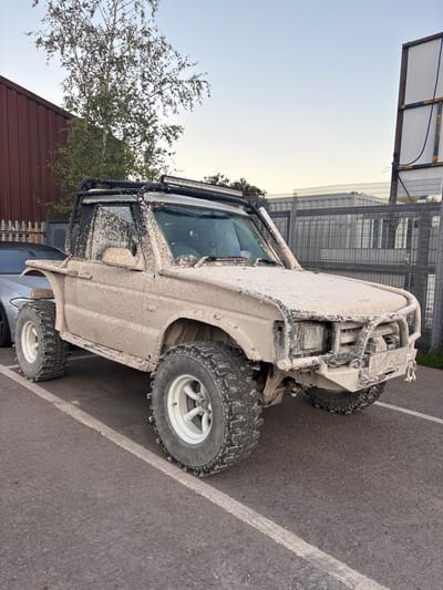 A heavily modified, mud-covered Land Rover Discovery, likely a Series I or II, is parked at an angle in an asphalt lot. The vehicle is completely caked in dried, light-brown soil, obscuring its original color. It features a black external roll cage, a roof-mounted LED light bar, a black snorkel on the passenger-side A-pillar, an aftermarket steel front bumper with recovery shackles, and large off-road tires on white multi-spoke alloy wheels. The rear bodywork is visibly shortened, with a wooden block wedged beneath the rear fender on the driver's side. A silver vehicle is partially visible parked immediately to the left. The background consists of a reddish-brown corrugated metal building and a leafy tree to the left, and a chain-link fence with barbed wire, grey shipping containers, and a tall white sign structure to the right. The sky is clear and light blue. No individuals are present, and no actions or interactions are depicted. The precise location within Nibley, United Kingdom, cannot be determined from the image, but the environment suggests an industrial or commercial area.