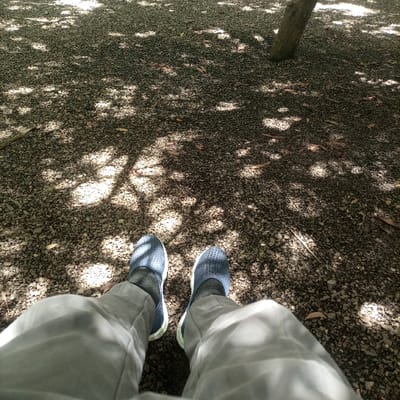 The image displays a first-person, downward view of an individual's lower legs and feet, located in Thika Township ward, Kenya. The person wears light-colored trousers, dark-colored socks, and dark blue, perforated slip-on shoes with white outer soles. The feet are positioned on a ground surface composed of small, irregular light-brown and grey gravel. Scattered dry, brown leaves and thin twigs are visible across the gravel. The environment is characterized by overhead foliage casting strong dappled sunlight and shadows, creating a mosaic pattern of light and dark areas on the ground. A segment of a tree trunk is visible in the upper right quadrant of the frame.