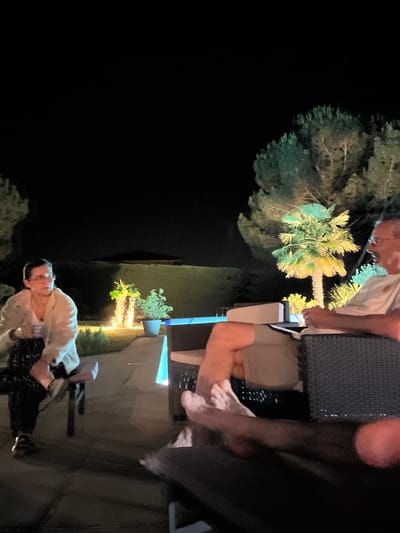 This is a vertical, medium-shot, eye-level photograph taken at night in the city of Saint-Galmier, France. The scene depicts a social gathering outdoors in a garden or patio area.
In the foreground on the left, a woman with short dark hair and glasses is sitting on a dark wooden bench. She is wearing a light-colored cardigan over a white top and patterned pants, with dark shoes. She is looking to her left, away from the camera.
In the middle ground, there are several elements. Two palm trees are illuminated by upward-facing lights, casting a warm glow. A potted plant sits to the right of the palm trees. A blue light source is visible in the far right middle ground, possibly a pool light or accent lighting.
On the right side of the frame, a man wearing glasses and a light-colored t-shirt is seated in a dark wicker chair, facing away from the camera and towards the left. His legs are crossed, and he is holding an object in his hands. His bare feet are visible in the lower right foreground, resting on what appears to be a dark cushioned seating area. The feet of at least one other person are also visible in the lower foreground, suggesting multiple people are present and relaxed.
The background is dark, indicating nighttime, with some trees visible against the dark sky. A subtle gradient of light suggests a distant landscape or elevation change. The overall atmosphere appears relaxed and informal.