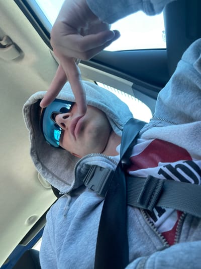 A person wearing a grey hoodie and blue sunglasses, secured with a seatbelt, is looking down at the camera while making a peace sign with their left hand. The photo appears to be taken from inside a vehicle. The location of Castrocielo, Italy, is not evident in the image.