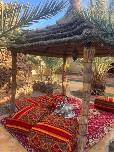 A shaded outdoor seating area displays colorful patterned cushions and a silver tea set on a rug. The seating is nestled beside a rock formation, with a thatched roof and palm fronds providing shade. A sandy pathway leads away from the seating, lined with palm trees and greenery, under a bright blue sky. The structure's materials and setting suggest a desert or arid climate.