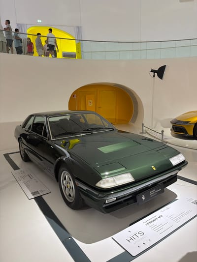 This image depicts a dark green Ferrari 412 parked indoors, likely in a museum or exhibition space. In the background, a large, curved, bright yellow archway is visible, suggesting a modern architectural design. To the right of the Ferrari, a portion of a yellow sports car is also visible. A spotlight is positioned on a stand above and to the right of the green Ferrari. In the upper left portion of the image, a group of people are standing on an elevated walkway, observing the exhibit. A railing separates them from the area below. A placard in front of the Ferrari provides information about the car, including its name, "Ferrari 412," and likely its origin or affiliation with "Museo Enzo Ferrari Modena." The overall environment appears to be a clean, well-lit indoor space designed to showcase automobiles.