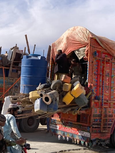 The image depicts an outdoor scene in Chaman City Tehsil, Pakistan, under a bright, partially cloudy sky. Two heavily overloaded trucks are the primary subjects.
The truck on the right is a red vehicle, extensively decorated with traditional Pakistani truck art in vibrant colors and intricate patterns. Its rear cargo area is covered by a large, peach-colored tarp, creating a tent-like structure. The rear bed is heavily loaded with diverse items, including multiple plastic jerry cans in yellow, blue, and orange, along with tires and miscellaneous bundled objects. A license plate displaying "TKA-464" and Urdu script is affixed to the lower rear of this truck. On the truck bed, near the tarp opening, a man in a dark jacket and light-colored trousers stands, with his hands positioned near his face. Partially visible inside the tarped enclosure, another man in a dark garment is seen holding a long, dark object.
To the left, the bed of a second truck is partially visible, carrying a large, blue cylindrical plastic water tank and numerous wooden poles and other bundled items.
In the lower left foreground, two men are seated on what appears to be a stationary motorcycle or scooter. The man in the foreground wears a light blue shalwar kameez and a white prayer cap. The ground beneath the vehicles consists of an uneven paved or dirt surface.