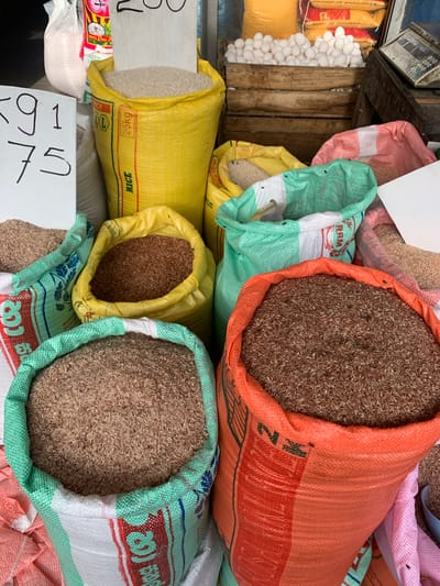 A market stall in Kodagoda, Sri Lanka, displays an assortment of large sacks filled with various types of rice. In the foreground, two open sacks are prominently visible: one is green with white and red accents, filled with light brown rice, and the other is orange with black and red markings, containing darker brown rice. Behind these, a yellow sack filled with white rice is partially visible, along with another green sack and a pinkish-red sack containing what appears to be grain. To the left, a white sign with handwritten black numbers "591 75" is propped up. In the background, a wooden crate overflows with white eggs, and a rustic weighing scale sits to the right. Several other sacks, some partially obscured, suggest a variety of products are for sale. The overall scene depicts a vibrant local marketplace.