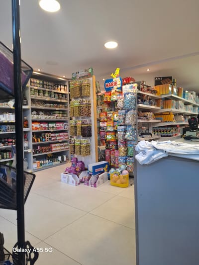 This media file captures the interior of a well-stocked retail shop, likely a convenience store or mini-market, located in Gouves Municipal Unit, Greece. The scene is brightly lit by recessed ceiling lights and shows multiple shelves filled with diverse merchandise.

In the upper part of the image, shelves display various packaged goods, including what appear to be books or board games, with one item clearly labeled "3-In-1." Below these, a large section is dedicated to sweets and candies, prominently featuring the "HARIBO" brand logo and its bear mascot on a large blue sign and numerous product packages. A yellow shopping basket rests on the light-colored tiled floor near this display.

Further down the shop, shelves are laden with packaged olives, indicated by a sign that reads "Greek Natural Olives." Below these, an array of jars and bottles suggests products like preserves, beverages, or alcoholic spirits. Other visible brands and text include "novela" and "Whiska." White clothing or textiles are hanging from a rack on the right side of the frame. A black metal railing, possibly part of a shopping cart or display unit, is visible in the foreground. No individuals are present in the shop. A "Galaxy A55 5G" watermark is seen in the bottom right corner of the image.