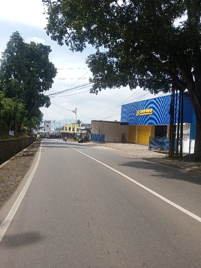 This image depicts a daytime scene along a paved roadway in Chhenangri_Rishor, Bhutan, under a partly cloudy sky. The perspective is from the roadside, looking down the two-lane asphalt road which features a solid white line on the right edge and a continuous white line marking the center.
On the left side of the road, a dirt shoulder with loose gravel adjoins a concrete gutter. Beyond the gutter, a low concrete retaining wall is visible, backed by dense green foliage and a line of trees extending into the distance. In the far background on this side, a light-colored vehicle, possibly a bus or van, is present on the road.
On the right side of the road, a substantial tree with dark green leaves occupies the immediate foreground, its branches extending over the road. Behind the tree, a large blue commercial building with horizontal corrugated siding features a prominent yellow sign displaying "DOWO BANGUNAN" in white text. To the left of this blue structure, additional single-story buildings are visible, some painted tan or yellow. Blue metal barrier fencing is positioned in front of these structures. Further down the road on the right, smaller buildings with varied paint schemes, including yellow and green sections, are present. Overhead, multiple power lines crisscross the frame.
Two motorcycles, each with a single rider, are observed traveling in the distance down the center of the road. Mountains are faintly visible on the horizon beneath the light blue, cloudy sky.