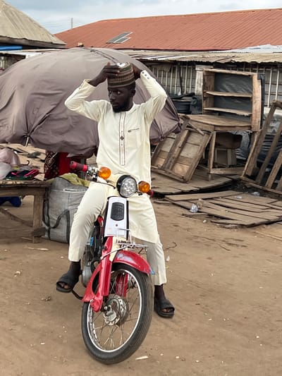 The image shows a man sitting on a red and white motorcycle in what appears to be an outdoor market in Oko Amakom, Nigeria. He is wearing a cream-colored traditional outfit and a striped cap, which he is adjusting. The motorcycle has chrome handlebars, orange turn signals, and a black seat. In the background, there are market stalls with tarpaulins, wooden structures, and various goods. The ground is dirt. The sky is overcast.