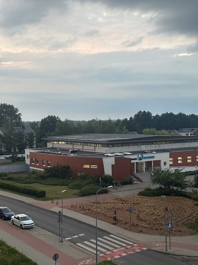 This is a high-angle, medium shot of a building in Żary, Poland, on an overcast day. The building, identified by the "WODNIK" logo, appears to be a public facility, possibly a sports center or aquatic facility, given the logo's wave-like design. The building has a modern architectural style with red brick accents and a white facade. There are cars parked along the street in front of the building, and a pedestrian crossing is visible. The surrounding area is green, with trees and landscaping. The sky is cloudy, and the lighting suggests it is either early morning or late evening.