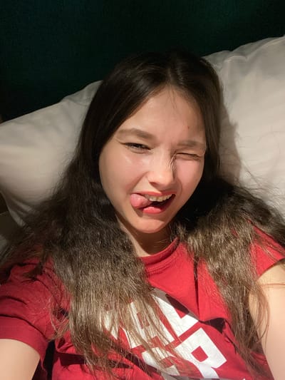 The image displays a playful indoor portrait of a young person, likely female, with long, dark brown wavy hair spread across a white pillow. The subject is wearing a red t-shirt, on which white letters are visible; the letter 'R' is clearly discernible, with other letters partially obscured. Lying down, the person makes a humorous face, sticking out their tongue, squinting or winking with one eye, and showing their teeth in a wide grin. The lighting suggests an indoor setting, possibly a bedroom. A dark green surface, likely a wall or blanket, is visible in the background on the left. The photograph, taken in Peresvet, Russia, conveys a lighthearted and relaxed mood.