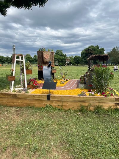 This detailed description pertains to an outdoor scene in Andrézieux-Bouthéon, France, featuring a prominently displayed raised garden bed in a large grassy field under an overcast sky.
The central subject is a rectangular raised garden bed constructed from horizontal wooden planks. Inside, the ground is predominantly covered with vibrant yellow petals or decorative chips. A narrow, reddish-brown pathway with two thin white lines, resembling a miniature running track, curves through the yellow ground covering on the right side of the bed.
On the far left of the garden bed, a white wooden ladder-like structure stands upright. Two brown rectangular planters are attached to it, containing small flowering plants with orange and purple blossoms. More red, purple, and white flowering plants are arranged at the base of this structure.
In the center, a large, cylindrical, bark-covered tree stump or log is positioned vertically, with several potted plants on its top surface. A circular, light brown target-like object is affixed to its front. Below this, a tall, black, rectangular reflective panel is angled downwards, reflecting the grey sky. A small, black, rectangular sign on a stand is placed in front of this panel.
Towards the right side of the garden bed, a small, open-sided structure with a thatched roof is visible. A large, leafy green shrub with numerous pink and red flowers grows prominently in front of this structure. To its left, a white tennis racket with red strings is partially visible, leaning vertically, alongside a yellow tennis ball. Another tennis racket is visible leaning against the thatched structure. Various flowering plants with red, white, pink, and yellow blossoms are dispersed throughout this section.
The garden bed is situated on a vast, light green grassy field that appears somewhat dry or patchy. A line of dense green trees forms the horizon in the background. The sky is completely covered with thick, grey clouds. In the distant background, to the right, two indistinct figures are visible walking on the grass. A dark green tree branch extends into the frame from the top left corner.