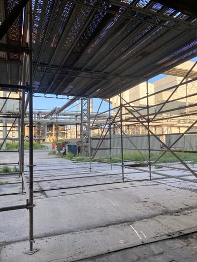 An outdoor industrial scene, likely a construction or demolition site, is depicted. The foreground is dominated by scaffolding structures, composed of metal poles and mesh platforms, creating an overhead canopy. The ground is a rough, concrete surface segmented by lines, with some areas of scattered debris and sparse vegetation. In the midground, a person wearing an orange vest walks away from the camera. Beyond them, a complex network of pipes and industrial buildings are visible under a clear blue sky. The overall impression is one of an active work environment within an industrial complex, possibly in or near the city of Police, Poland, given the context of the prompt.
