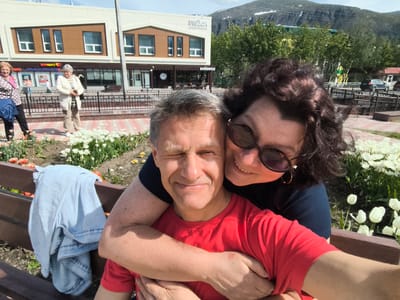 A man and a woman are posing for a selfie in Kirovsk, Russia. The man is in the foreground, wearing a red t-shirt. The woman is behind him, embracing him and wearing a dark blue top and sunglasses. They are sitting on a wooden bench, with a flowerbed of white tulips to their right. In the background, there is a modern building with a sign that appears to be in Russian. The building is flanked by trees, and beyond them, a mountainous landscape with patches of snow is visible. Other people and parked cars can be seen further in the background. The overall impression is a candid, outdoor moment in a pleasant, scenic environment.