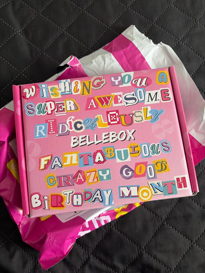 A pink cardboard box with vibrant, magazine-cutout-style lettering is featured prominently, resting on a dark, quilted surface. The box, partially removed from its white and pink plastic mailing bag, displays the text: "WISHING YOU A SUPER AWESOME RADICAL BELLEBOX FABULOUS GOOD CRAZY BIRTHDAY MONTH." The mailing bag, torn open, reveals a small, partially obscured text in Bulgarian: "ЕКЗЕМПЛЯР НА ПОЧ", likely part of "ЕКЗЕМПЛЯР НА ПОЩАТА" (copy for the post office), suggesting the package was sent via post. The image captures the moment of receiving or unboxing a package, likely a subscription box given the "BELLEBOX" branding. The quilted background suggests an indoor, domestic setting. The package was received in Novi Pazar, Bulgaria.