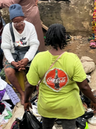 Two individuals are positioned amidst a collection of miscellaneous items. The person on the left wears a white long-sleeved shirt with a black belt, a blue knitted cap, and spectacles. The person on the right wears a lime-green short-sleeved shirt adorned with a Coca-Cola logo. They clasp each other's hands, the surroundings cluttered with clothing, bags, and other objects scattered about. The setting appears to be Idimu, Nigeria.