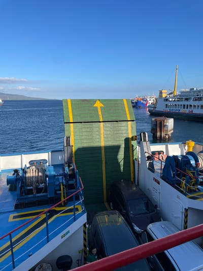 The image depicts the deck of a ferry carrying vehicles, likely at a port. In the foreground, a green ramp with a yellow arrow points upwards, indicating the direction of travel. Several vehicles, including a dark SUV and a lighter car, are parked on the deck. Two large winches with ropes are visible on the left side of the ferry. In the background, other ferries are docked or sailing in the water. The sky is clear and blue, and a distant landmass with hills can be seen on the horizon. The overall scene suggests a transportation hub, possibly a ferry terminal in Ketapang, Indonesia, given the context provided.
