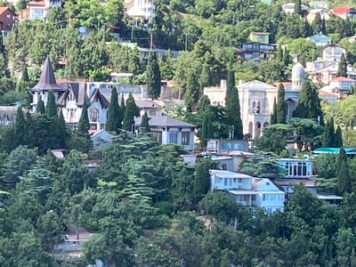 The image shows a view of several buildings nestled within a lush, green, hilly landscape. The architecture varies, with some buildings exhibiting a more traditional or gothic style, featuring pointed roofs and decorative elements, while others are modern with flat roofs and large windows. The buildings are surrounded by dense evergreen trees, suggesting a temperate climate with significant vegetation. Some structures appear to be residential, with visible balconies and windows. The overall impression is a scenic, perhaps secluded, community situated on a slope. The specific location within Симеизский поселковый совет, Ukraine, is not explicitly identifiable from the image itself, but the presence of diverse architectural styles and the natural environment are the key visual elements. There are no visible people or discernible actions taking place in the scene.
