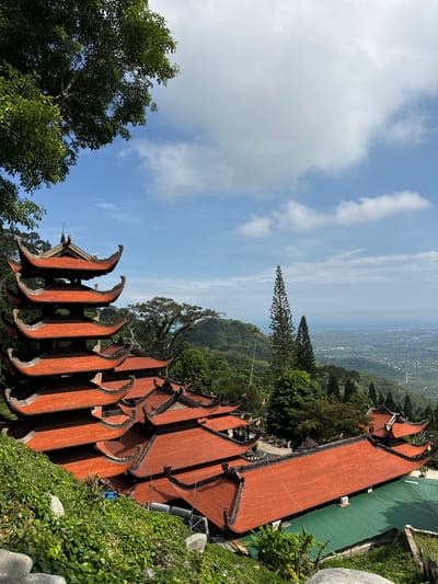 The image captures a panoramic view of a temple complex with distinct, ornate red-tiled roofs, set against a lush, mountainous landscape. The traditional architecture, characterized by curved eaves and detailed embellishments, suggests a location in Southeast Asia, consistent with the provided context of Thuận Nam, Vietnam. The temple appears to be situated on a hillside, offering a sweeping vista of a town or village nestled in the valley below, which stretches towards a distant body of water.

The sky above is a bright blue, punctuated by fluffy white clouds, indicating fair weather and likely daytime. The sunlight casts shadows, highlighting the textures of the tiled roofs and the dense green foliage surrounding the structures. A tall, slender evergreen tree stands prominently on the slope, its unique shape adding to the visual interest of the scene.

The image is taken from a high vantage point, looking down and across the temple buildings and the expansive landscape. There are no visible people in the frame, nor any explicit signs of an ongoing event. The focus is on the serene beauty of the temple architecture and its harmonious integration with the natural environment. The overall mood conveyed is one of peace and grandeur, emphasizing the spiritual and aesthetic qualities of the location. The presence of modern elements like air conditioning units attached to the temple walls provides a subtle contrast to the traditional design.