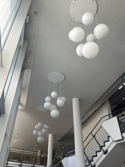 This image captures a modern interior space with a high ceiling and a large window on the left. The ceiling features recessed lighting and multiple contemporary pendant light fixtures composed of white spherical globes. In the foreground, a white cylindrical pillar stands prominently. To the right, a staircase with a black metal railing and wooden handrail ascends. The overall environment suggests a public building, possibly a library or a corporate office, located in Ludwigsburg, Germany. There are no people visible in the frame.