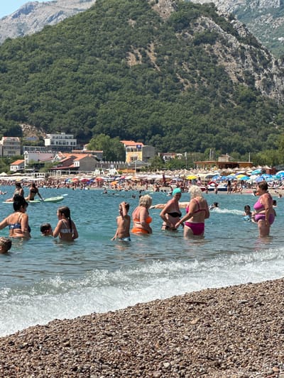 The image shows a crowded beach in Đurovići, Montenegro. In the foreground, several people are standing in the shallow water. To the left, a woman with dark hair in a blue and white bikini is partially submerged, with a child visible to her left. Beside her, a young girl with her hair in pigtails wears a patterned bikini, and a boy with dark hair and a red headband stands next to her, facing left. Further to the right, a man with dark hair and light blue shorts is also in the water, looking towards the right. In the middle ground, a woman in an orange bikini is visible, and to her right, a woman in a black bikini with a green headscarf is speaking to a blonde woman in a pink bikini. Behind them, other beachgoers are in the water and on the shore. The beach is lined with numerous colorful umbrellas, and buildings are visible in the background, nestled against a lush green hillside that rises to rocky mountains. The sea is calm, with small waves breaking on the pebble beach.