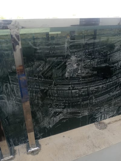The image shows a close-up view of a dark, textured surface that appears to be a glass panel with a protective film or coating. The surface is marred with numerous streaks, smudges, and peeling sections of this film, creating an uneven and abstract pattern. On the left side of the image, a vertical, polished metal post is visible, attached to the panel with square, silver bases secured to a concrete surface below. The concrete surface is light-colored and appears weathered, with some small cracks and patches of discolored material. In the upper portion of the image, a thin, horizontal metal band runs along the top edge of the glass panel, with faint text indicating "SUS 304 Made in Turkey." The background is blurred, showing reflections of what could be buildings and a fence. The overall scene suggests an outdoor setting, possibly a railing or a barrier, in a state of disrepair or undergoing installation/removal of protective material. The location in Hotoro North, Nigeria, is not visually evident from the provided image.