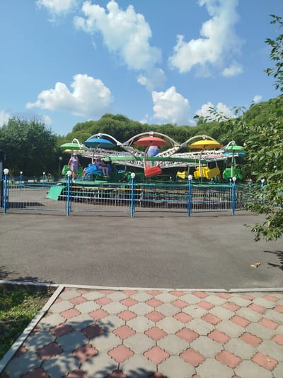 An amusement ride with multiple colorful cars, each with its own canopy, is set up in a park under a partly cloudy blue sky. The ride features green and blue fencing around it, with a paved walkway in the foreground. Two individuals are visible on the ride, one standing and one seated in a car. Trees and foliage form a green backdrop. The image was taken in the city of Yalutorovsk, Russia.