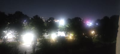 This image captures a nighttime scene in Andijan City, Uzbekistan, likely from an elevated viewpoint looking out over an area with trees and buildings. The sky is dark, suggesting late evening or early morning. Several bright lights illuminate the scene, some appearing to be streetlights or lights from buildings. These lights cause significant glare and diffusion, obscuring details in the immediate foreground and midground. Trees are prominent, silhouetted against the brighter areas, with some foliage visible where it is caught by the light. In the distance, buildings with illuminated windows are visible, and there are also indications of distant lights with a pinkish hue, possibly from signage or illuminated structures. The ground appears wet or reflective in some areas, suggesting recent rain or dampness. There are no clear indications of people or specific activities taking place. The overall impression is a somewhat hazy and atmospheric urban landscape at night.
