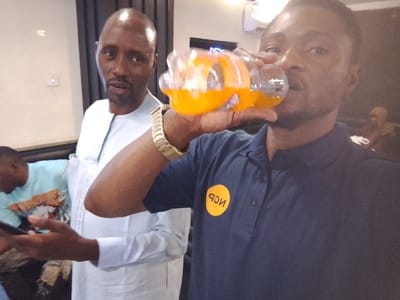 Two men are pictured indoors. One, in the foreground, is drinking orange juice from a glass bottle, wearing a navy blue polo shirt with a round, yellow logo. A gold wristwatch is on his wrist. Behind him is a man in a light-colored, formal shirt. Others are visible in the background. The lighting is soft, and a dark curtain is partially visible behind the standing men.