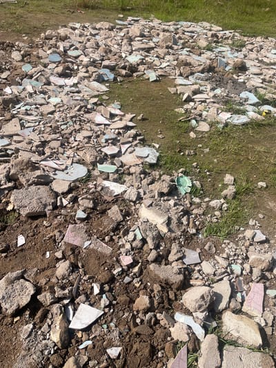 A pile of rubble, consisting of broken concrete chunks, tiles, and dirt, is scattered across a patch of muddy ground with sparse green grass. The rubble is a mixture of light brown, grey, and some pieces with remnants of pink and blue glaze, suggesting they were once decorative tiles. The scene appears to be an outdoor area, possibly a construction or demolition site, within Wauro Jabbe, Nigeria. There are no people or animals visible in the image. The lighting suggests it is daytime, with sunlight casting shadows on the debris.