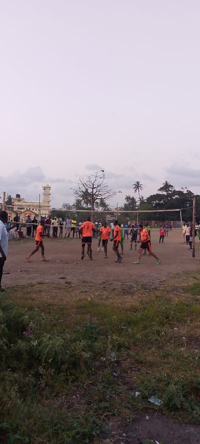 The image displays an outdoor volleyball game in progress on a dirt court in Magogoni ward, Kenya. Approximately 10-12 male individuals are actively participating in the game. Several players wear matching orange jerseys and dark shorts; one player facing away from the camera exhibits the number 6 on his jersey. Other players are observed in various athletic wear, including green, blue, and black attire, positioned dynamically on the court. A man in a light blue shirt is partially visible on the far left, observing the activity. A crowd of spectators lines the court's perimeter.
The playing surface consists of packed dirt with sparse patches of dry grass and weeds in the foreground. A white volleyball net is stretched across the court, supported by two wooden posts.
In the background, a multi-story building with yellow and white paint featuring a prominent tower topped with a golden dome is visible to the left. Further right, additional buildings, a large deciduous tree, and several palm trees are present under an overcast, light grey sky.
