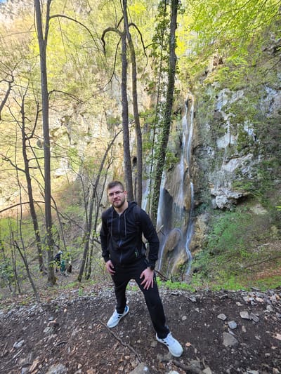 A vertical shot shows a Caucasian man with short brown hair and a beard standing in a wooded area at the Bachkovo Monastery in Bulgaria. He wears glasses, a black tracksuit, and white sneakers. He faces the camera with his hands on his hips. The ground is a mix of soil and rocks. In the background, a waterfall cascades down a rocky cliff face. Trees with budding green leaves surround the area.