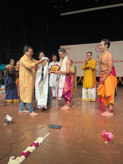 The image shows an award ceremony on a stage in Chandigarh, India, at the location with coordinates 30.733 latitude, 76.789 longitude.  A man in a yellow kurta is presenting a white shawl to another man wearing a white dhoti and a pink dupatta. The man receiving the shawl is holding a plaque.  There are three other men standing on the stage behind the recipients, and two women are seated to the left.  A white garland of flowers lies on the wooden floor, with a black object and two small red and white objects on the floor.  The background includes a banner with text in Hindi and English.  The stage is lit with artificial lighting.