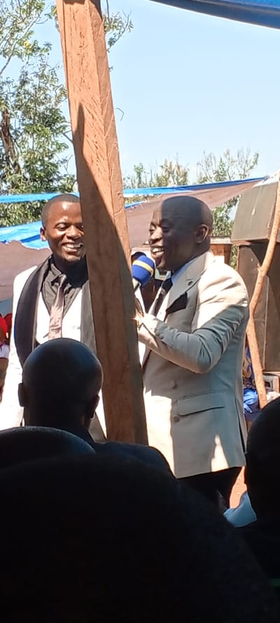 The image shows two African men outdoors, possibly in Kalemie, Democratic Republic of the Congo, based on the context of the query. One man, on the left, is wearing a white suit with a black tie and a dark scarf draped over his shoulders, he is smiling. The other man is on the right, wearing a light-colored suit and holding a microphone with a blue windscreen. Both men are smiling and appear to be engaged in a positive interaction. A wooden post is positioned prominently in the foreground between the two men. In the foreground, the backs of the heads of individuals in the audience are visible, appearing as dark shapes. The background includes trees and a light-colored canopy. The lighting suggests it is daytime.