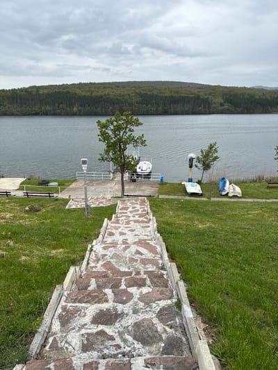 The image shows a view of a lake surrounded by forest, likely in the Rakilovtsi, Bulgaria area, as indicated. The view is from an elevated position, looking down a stone and concrete staircase. To the right of the staircase is a green grassy area, and beyond that, the lake. On the lake's edge, there are several boats, including a pedal boat and other small watercraft. There's also a weather station-like structure next to a mature tree. The sky is overcast, suggesting a cloudy day.