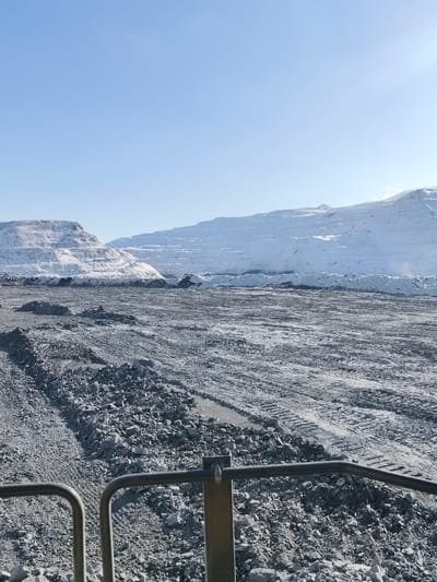 The image depicts an open-pit mining operation in the city of Im. Gastello, Russia, under clear daylight conditions. The foreground consists of rough, disturbed grey ground composed of angular rocks and gravel, displaying multiple tire tracks from heavy machinery. In the immediate bottom center, two dark grey metal railings and a partial yellow-brown vertical support are visible, indicating the perspective is from a vehicle or elevated platform. The midground extends as a vast, uneven expanse of similar rocky terrain, featuring scattered dark piles of excavated material. The background is dominated by large, snow-covered terraced slopes characteristic of open-pit mining, forming artificial mountains. At the base of these snow-dusted slopes, several dark-colored heavy mining vehicles, including what appears to be an excavator, are visible, engaged in excavation or material handling operations against piles of dark material. The sky above is clear and light blue, with no visible clouds. No individuals are distinctly discernible.