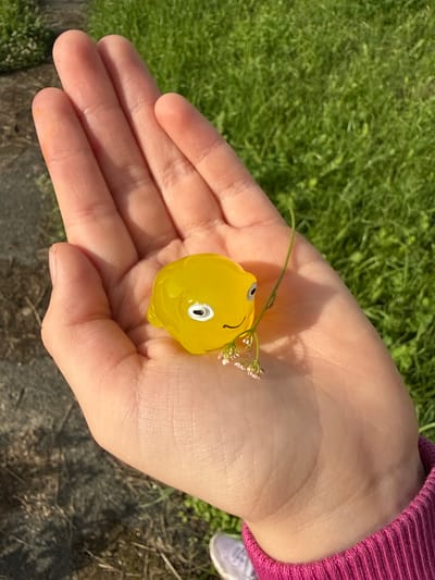A close-up shot shows a child's open palm holding a small, yellow, toy lemon with a cartoon face. The toy lemon has a white eye with a black pupil and a smiling mouth. A delicate green stem with tiny pink flowers is attached to the toy, arching over the lemon's face. The child's hand is pale and appears to be resting on a surface of green grass. The background is slightly blurred, but the texture of the grass and hints of dark soil are visible. The child is wearing a bright pink, ribbed long-sleeved shirt, and a small portion of a white sneaker with grey accents can be seen near the edge of the frame, suggesting the child is outdoors. The lighting indicates it is daytime, likely sunny, with distinct shadows cast on the hand. The overall scene evokes a sense of simple joy and discovery, possibly from a park or a backyard in Большой Турыш, Russia.