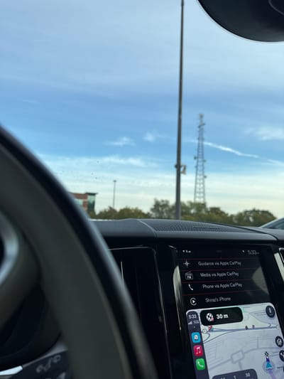 The image captures a scene from inside a vehicle looking through a smudged windshield. A portion of a beige/grey steering wheel is visible on the left, with the vehicle's black dashboard and infotainment screen below. The infotainment screen displays an Apple CarPlay interface, showing the time as 5:33 PM, connectivity options (Guidance, Media, Phone) via Apple CarPlay, and an active connection to "Shiraz's iPhone." A navigation map, partially visible on the lower screen, indicates a next turn or destination at "30 m" and displays "James Forrestal Road," confirming the location in Plainsboro Township, New Jersey, United States. The phone's status indicates a 5G signal and 50% battery.

Beyond the windshield, under a blue sky with scattered white clouds and a faint contrail, a tall street light pole stands prominently in the center. Further in the background and slightly to the right of the pole, a large metal lattice telecommunications tower is visible. A line of green trees separates the sky from the lower ground elements. To the far left, a multi-story building with dark paneling and lighter sections is partially visible. The scene is consistent with a commercial or office park environment within Plainsboro Township, New Jersey.
