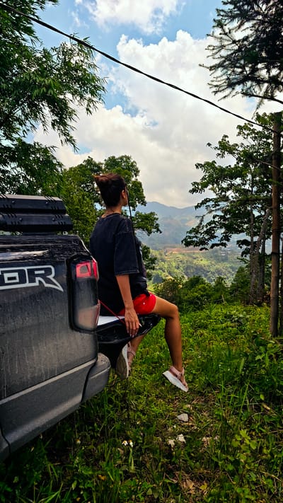 A woman with a top bun sits on the open tailgate of a dirty, black truck with the word "RAPTOR" on the side. She wears a dark, oversized t-shirt, red shorts, and light pink sandals. Her back faces the camera as she gazes at a hilly landscape in Hoàng Su Phì Commune, Vietnam. Green trees and vegetation surround the truck. Overhead electrical wires stretch across a blue sky with scattered white clouds. Tall grass and small white flowers grow around the truck. Wooden posts border the right side of the scene.